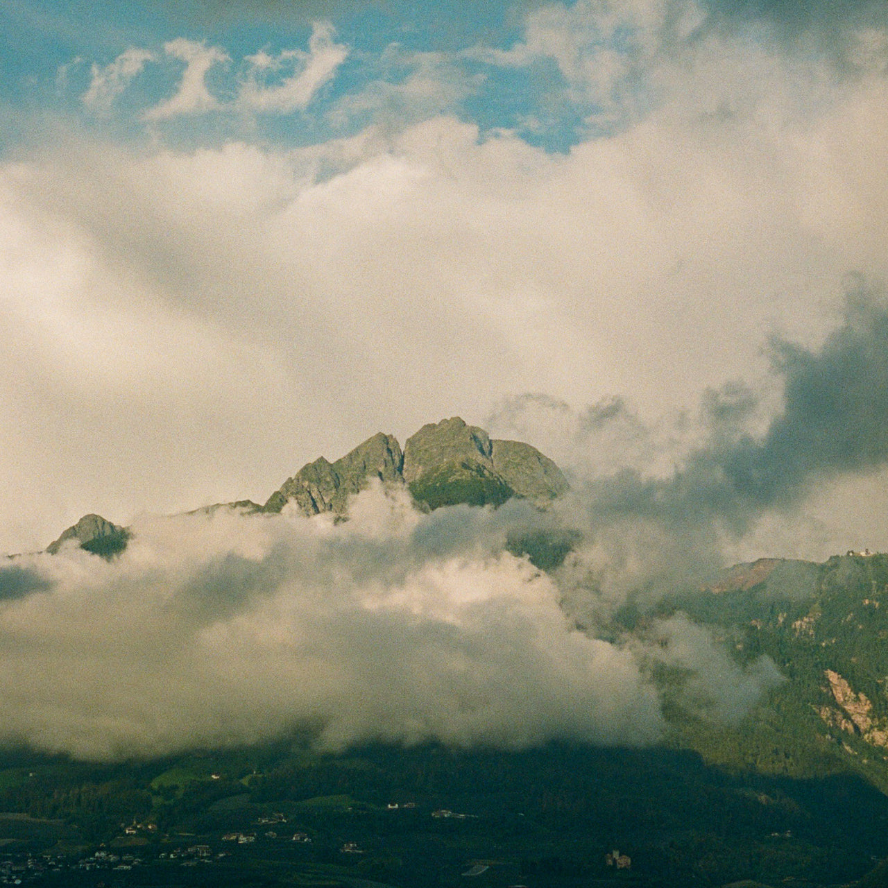 Eine dramatische Berglandschaft mit majestätischen Gipfeln, die teilweise von dichten weißen und grauen Wolken umhüllt sind. Der Himmel zeigt eine Mischung aus bläulichen und cremefarbenen Tönen, während im Vordergrund ein dunkles Tal mit grüner Vegetation zu sehen ist. Die Atmosphäre wirkt mystisch und stimmungsvoll.