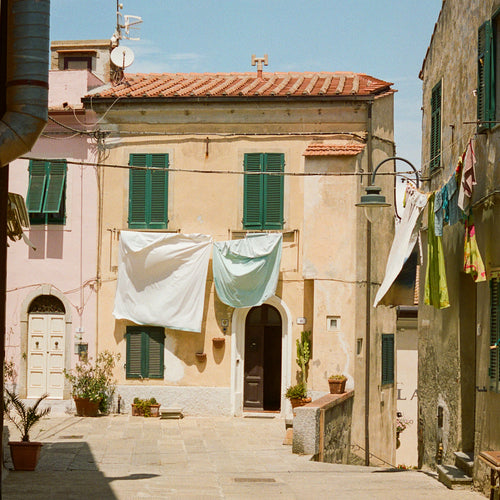 Eine malerische mediterrane Straßenszene mit traditionellen Häusern in beigen und cremefarbenen Tönen, grünen Fensterläden und roten Ziegeldächern. Zwischen den Gebäuden hängt Wäsche an Leinen - weiße Laken und bunte Kleidungsstücke. Auf dem gepflasterten Platz stehen Blumentöpfe mit Pflanzen, und im Hintergrund ist ein klarer blauer Himmel zu sehen.