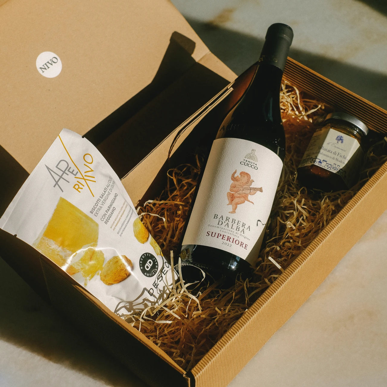 Ein Geschenkkarton aus Wellpappe mit braunem Füllmaterial, der eine Flasche Barbera d'Alba Superiore Rotwein, ein Paket OLIVO-Produkte mit gelben Lebensmitteln und ein kleines Konservenglas enthält. Der Karton steht auf einer Holzoberfläche und trägt ein 