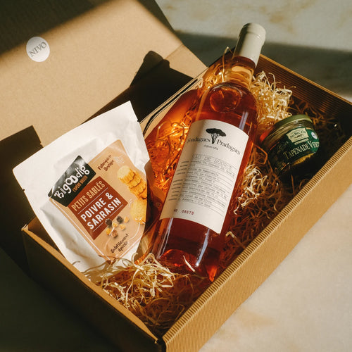 Ein Geschenkkarton aus Wellpappe mit braunem Füllmaterial, der eine Flasche Roséwein "Eau de Source" mit weißem Etikett, eine Packung Bruschette-Snacks "Petits Salés Poivre & Sarrasin" und ein Glas Tapenade enthält. Der Karton trägt ein "NIVO"-Etikett und steht auf einer hellen Oberfläche.