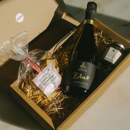 Ein Geschenkkarton aus Wellpappe mit braunem Füllmaterial, der eine Flasche Brut Schaumwein der Marke Tenuta Cucco, eine Packung Quadrini-Gebäck in durchsichtigem Beutel mit kariertem Etikett und ein kleines Konservenglas enthält. Der Karton trägt ein "NIVO"-Etikett und steht auf einer hellen Oberfläche.