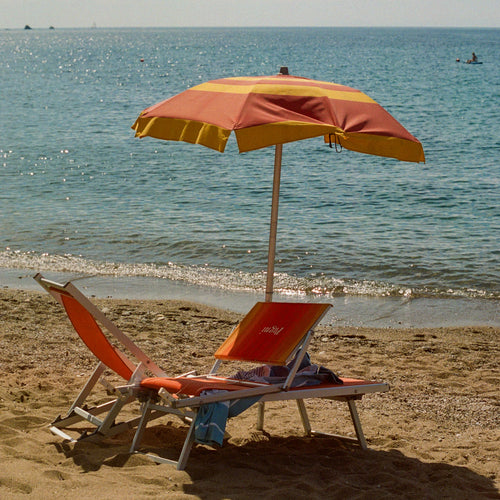 Ein orangefarbener Sonnenschirm und Liegestuhl am Sandstrand mit ruhigem blauem Meer im Hintergrund. Auf dem Liegestuhl liegen persönliche Gegenstände, und in der Ferne sind kleine Boote auf dem Wasser zu sehen. Die Szene vermittelt eine entspannte Urlaubsatmosphäre am Mittelmeer.