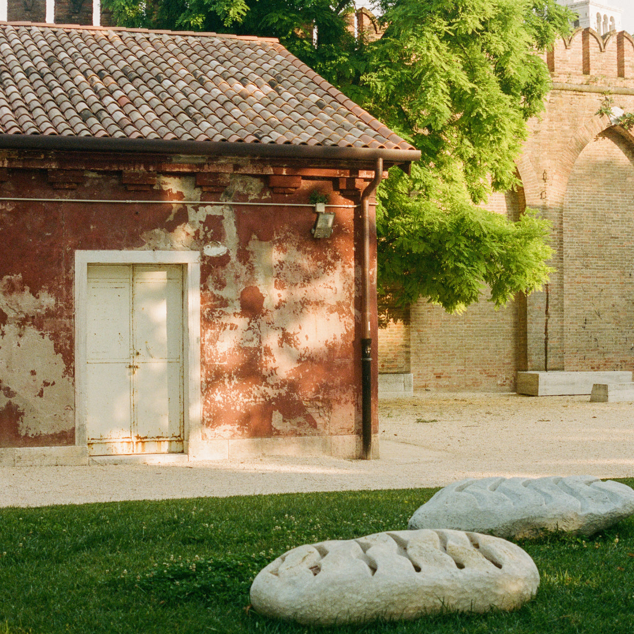 Ein rustikales Gebäude mit verwitterten rot-braunen Wänden und traditionellem Ziegeldach, vor dem eine weiße Holztür steht. Im Hintergrund ist historische Steinarchitektur mit Bögen zu sehen, umgeben von üppigem grünem Laub. Im Vordergrund liegen auf einer Rasenfläche mehrere geschnitzte Steinelemente oder archäologische Fragmente.