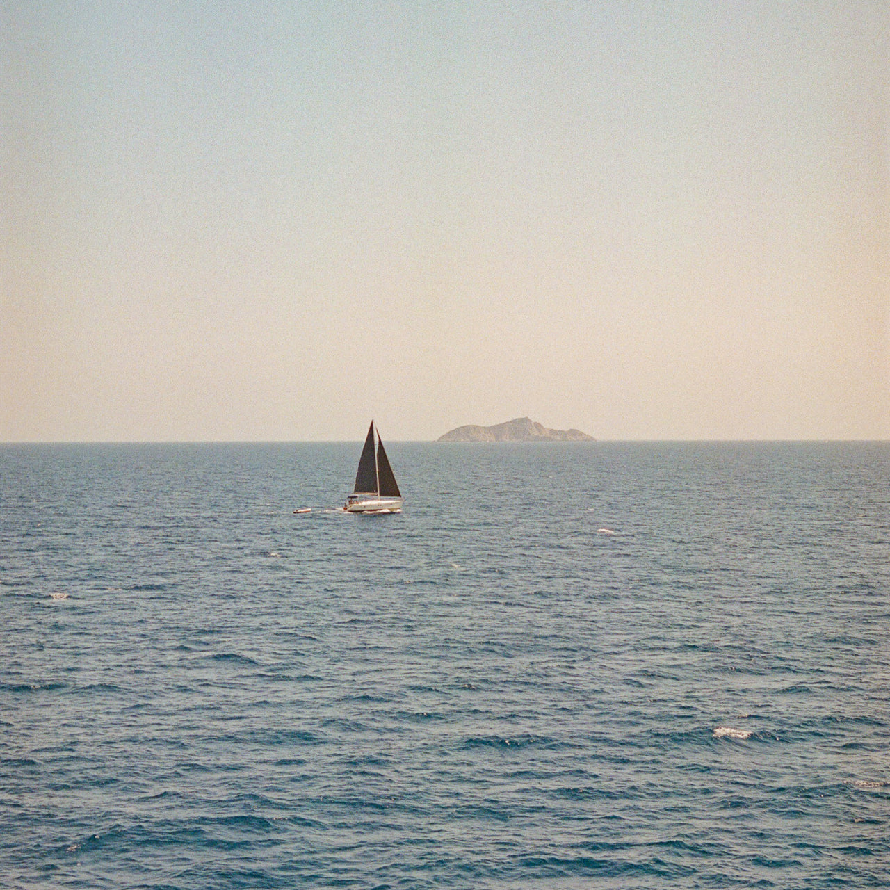 Ein Segelboot mit dunklen Segeln auf ruhigem blauem Meer. Am Horizont ist eine kleine Insel oder Landmasse zu sehen, während der Himmel in gedämpften grau-weißen Tönen erscheint. Die Szene vermittelt eine friedliche, maritime Atmosphäre.
