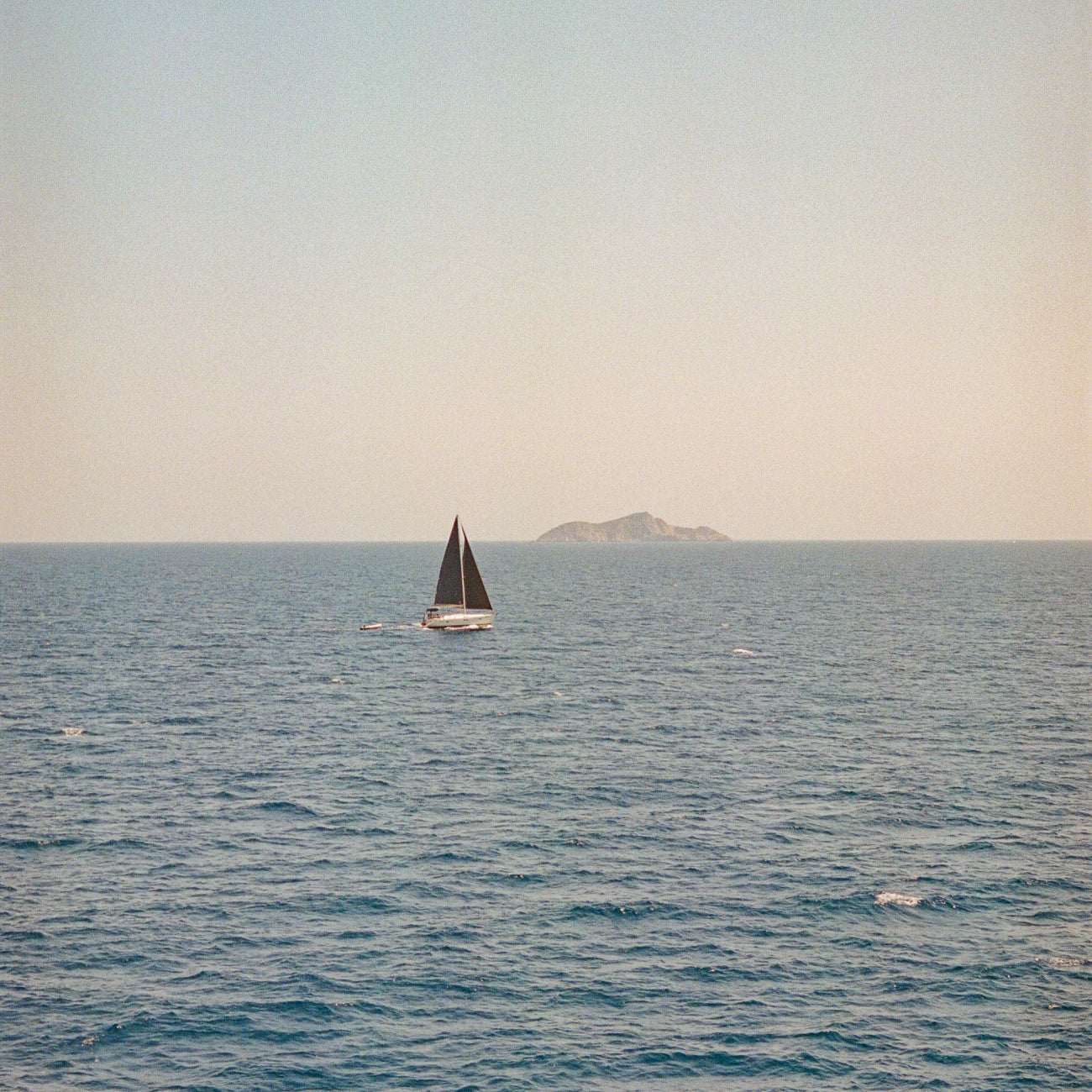 Ein Segelboot mit dunklen Segeln auf ruhigem blauem Meer. Am Horizont ist eine kleine Insel oder Landmasse zu sehen, während der Himmel in gedämpften grau-weißen Tönen erscheint. Die Szene vermittelt eine friedliche, maritime Atmosphäre.