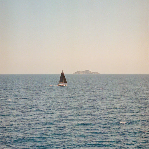 Ein Segelboot mit dunklen Segeln auf ruhigem blauem Meer. Am Horizont ist eine kleine Insel oder Landmasse zu sehen, während der Himmel in gedämpften grau-weißen Tönen erscheint. Die Szene vermittelt eine friedliche, maritime Atmosphäre.