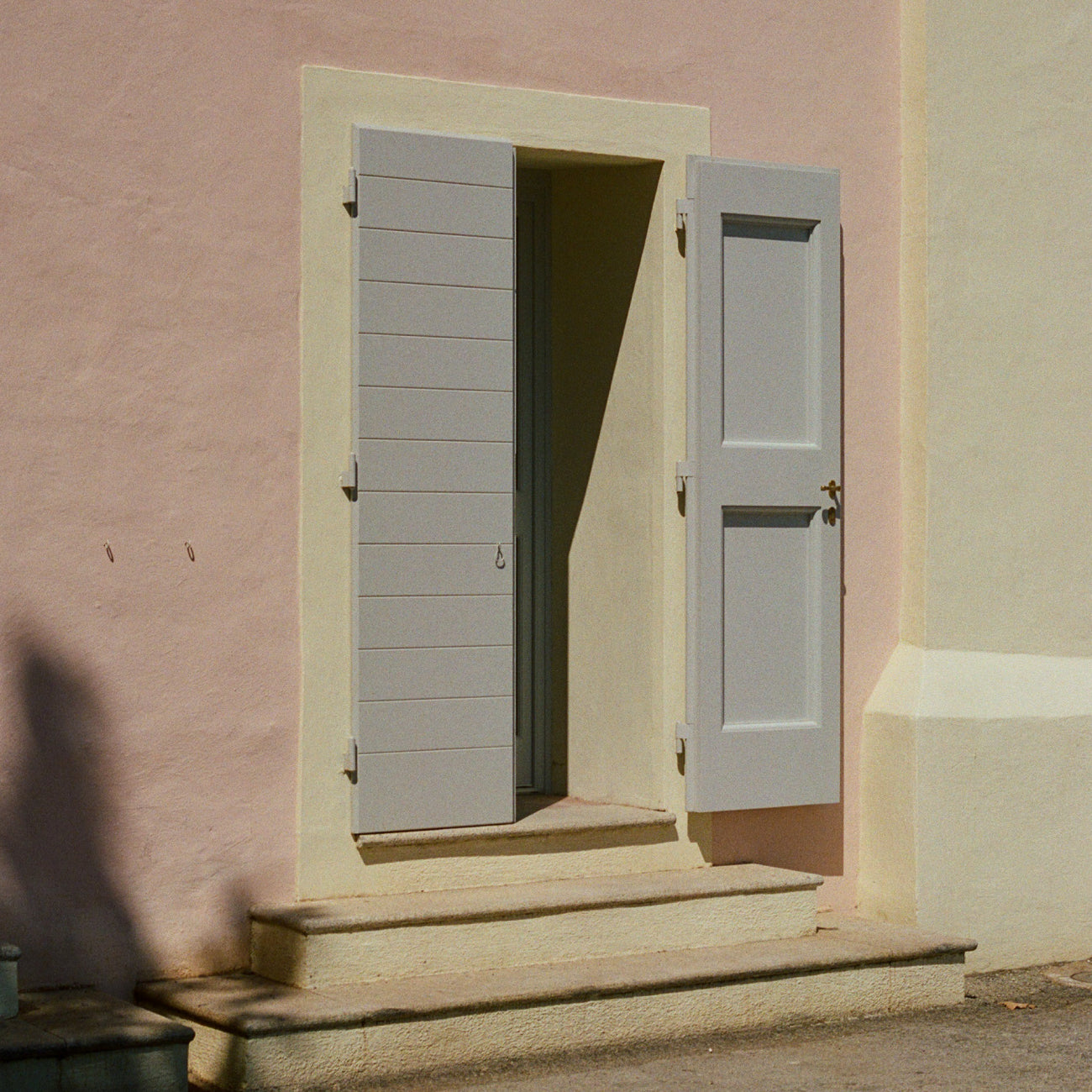 Ein Hauseingang mit rosa-beigen Wänden und einer offenen Tür, die von grauen Klappläden mit horizontalen Lamellen eingerahmt wird. Steinstufen führen zum Eingang hinauf, und warmes Sonnenlicht wirft Schatten an der Wand. Die Architektur zeigt einen typisch mediterranen Stil.